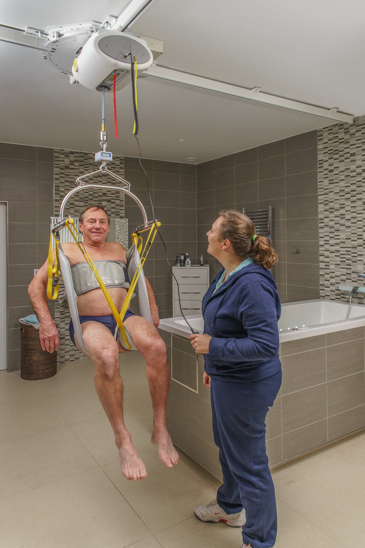 Veilige transfer van rolstoel naar bad met een plafondlift en tilmat in de badkamer.
